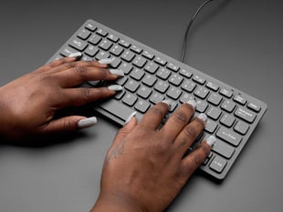Angled shot of a Black woman's silver-blue manicured hands on a slim, black keyboard.