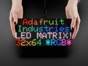 Two white hands hold out an assembled and powered on 64x32 RGB LED Matrix Panel - 4mm pitch. The matrix displays "Adafruit Industries LED MATRIX! 32x64 *RGB*"