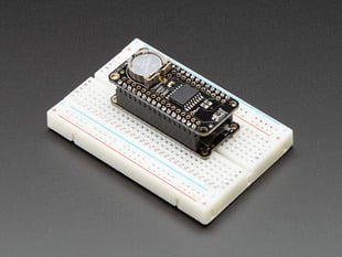Angled shot of a DS3231 Precision RTC FeatherWing - RTC Add-on For Feather Boards stacked on a rectangular breakout board, connected to a white breadboard. 