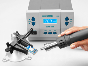 Lifestyle image of a hand holding a hot air rework nozzle onto a PCB in a vise. Hot air rework station powered on in the background.