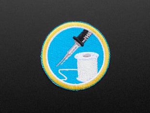 Circular embroidered badge with image of soldering iron and solder spool on blue background with yellow edge. 