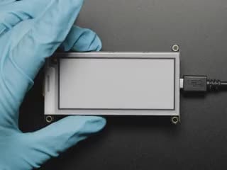 Demo Shot of the ePaper Display connected to a Feather as a Hand wearing a blue glove presses a reset button that make the display flash black and white a few times until the Adabot Character appears on the screen.