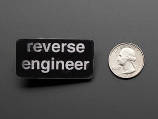 straight down view of black rectangular badge with the words REVERSE ENGINEER in white, next to a quarter for scale. 