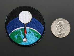 Circular badge with white high altitude balloon over a green and blue earth on a black background, with black trim. Shown next to a quarter for scale. 