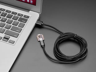 Angled shot of 2 meter long black, woven cable with USB-A and USB-C connectors connected to a laptop and a small, square microcontroller.