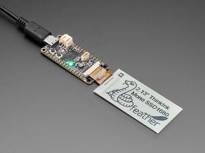Angled shot of a 2.13" monochromatic electronic ink display module connected to a driver board. The display features a graphic python with the text: 2.13" ThinkInk Mono SSD1680 feather"